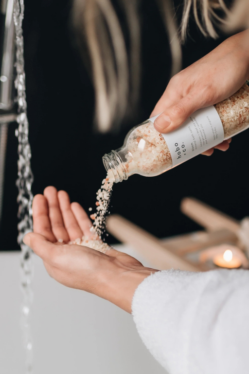 pink and white bath salts being poured into a womans hand in front of a running bath tap