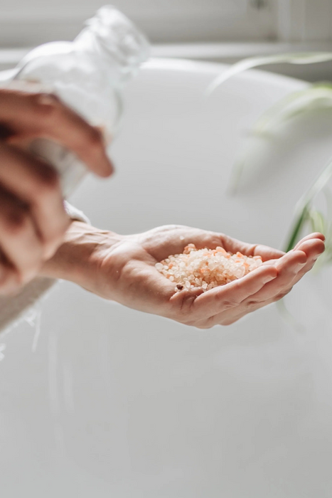 Pink and white bath salts being held in someones hand over a white bath, the glass bottle container for them being held in the foreground 
