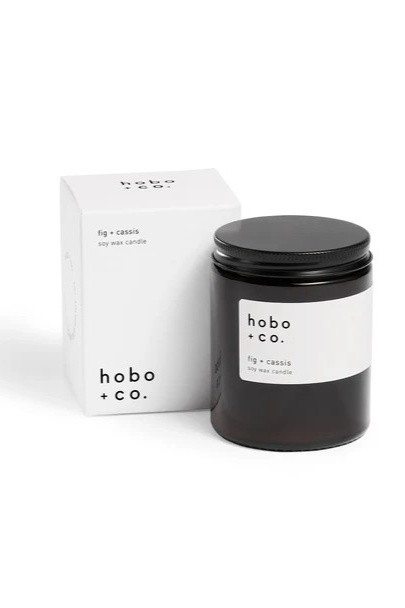 Dark glass jar candle with black metal lid with a white Hobo + Co. branded label, displayed with its its white card branded box