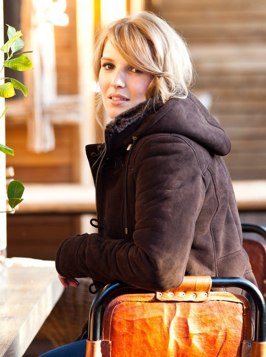 Millie Sheepskin Duffle Coat. Female model wears brown Sheepskin coat outdoors, sat on a leather seat